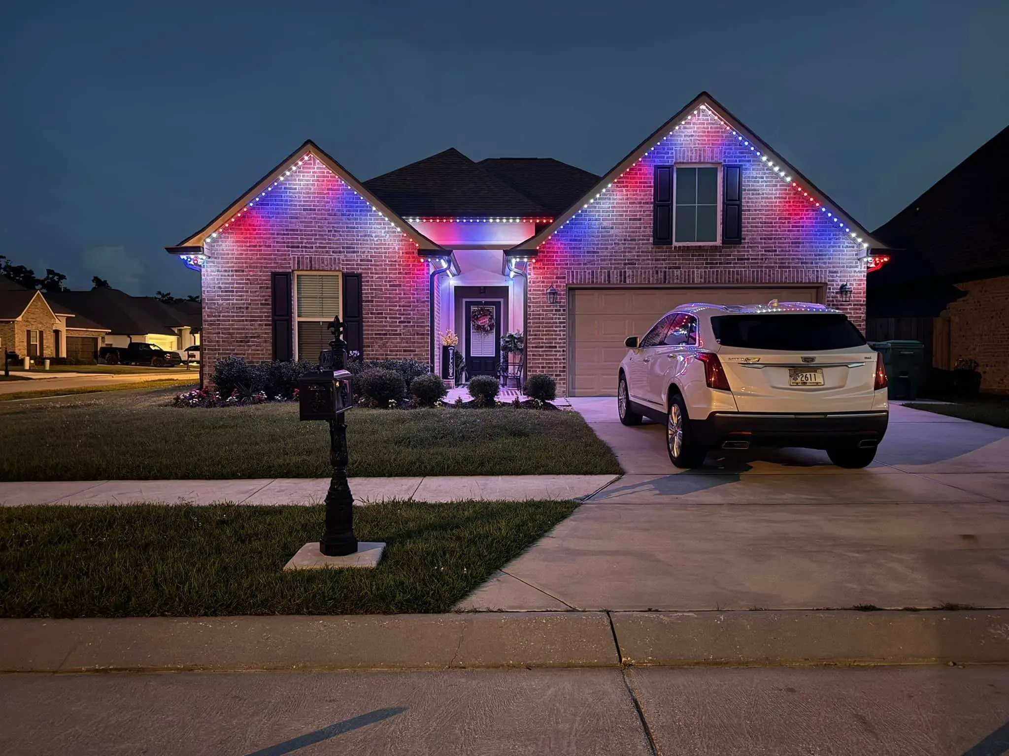Brick house at night with red and blue Trimlight along the roof and a white SUV in the driveway.